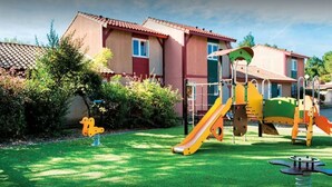 A playground with a slide and climbing structures, surrounded by a well-maintained lawn and a house with a red roof.