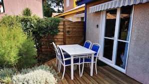 A patio with a white table and blue chairs, surrounded by a wooden deck and various plants.