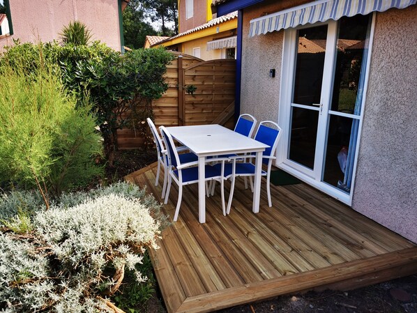 Un patio con mesa blanca y sillas azules, rodeado por una terraza de madera y diversas plantas.