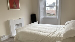 Een slaapkamer met een wit bed, een witte radiator, een raam met uitzicht op gebouwen en een ingelijst schilderij aan de muur.