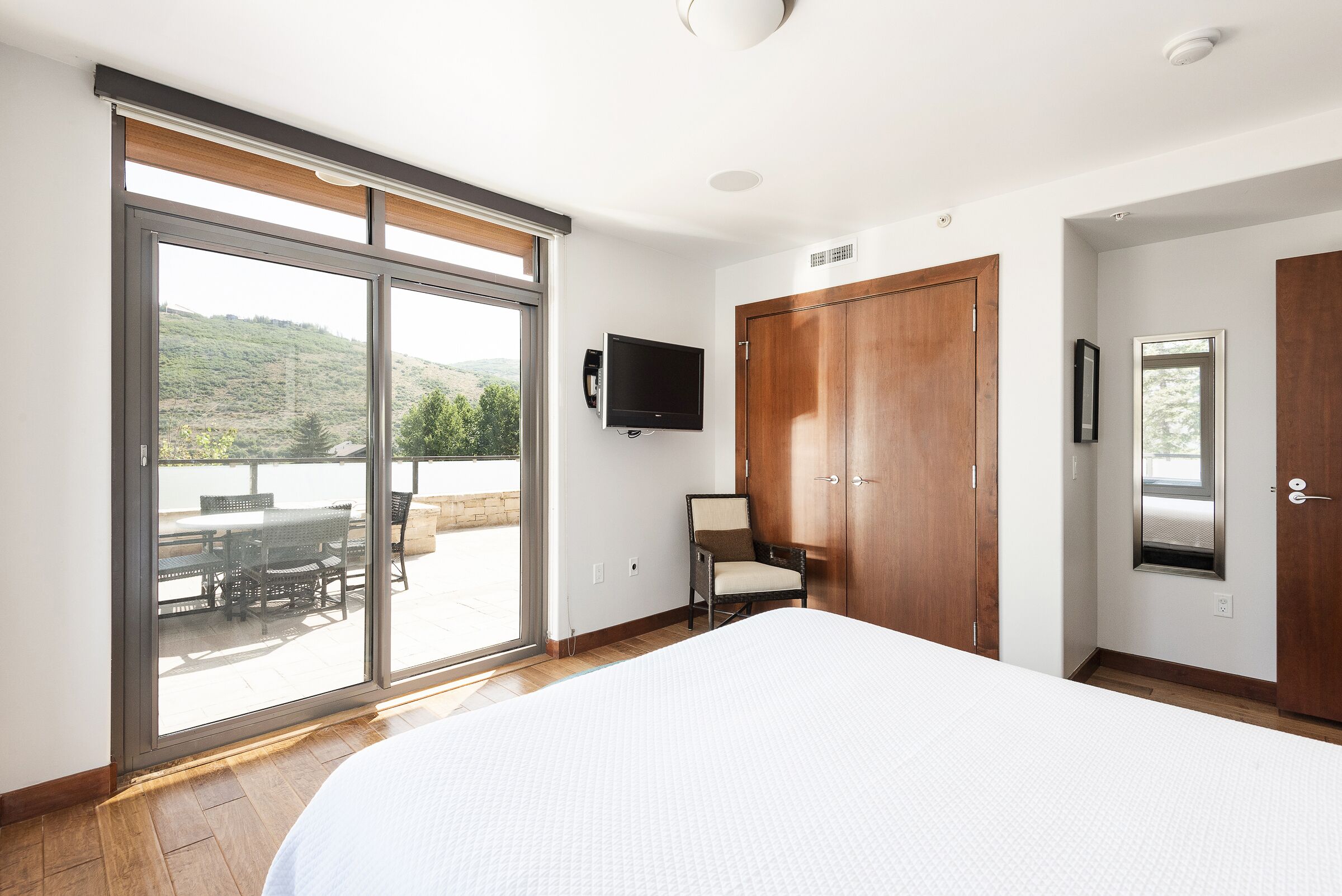 Une chambre avec un grand lit, une porte-fenêtre coulissante donnant sur une terrasse, un téléviseur fixé au mur et des armoires en bois.