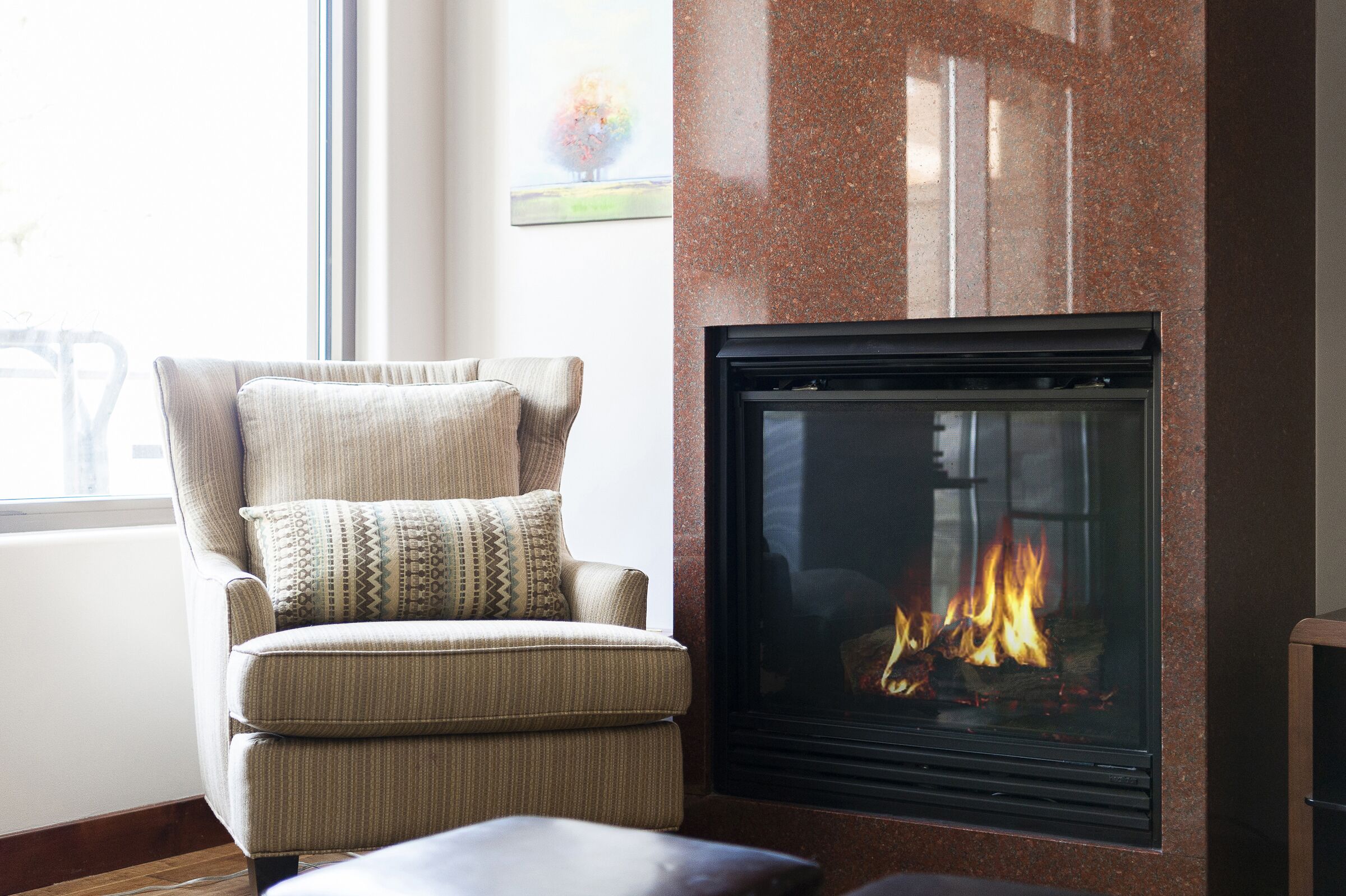 A living room with a fireplace, a comfortable armchair, and a view of the outdoors through a large window.