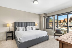 A modern bedroom with a large bed, two bedside tables, a chair, and a view of a building complex.