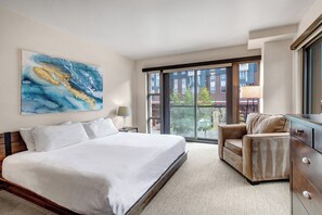 A hotel room with a large bed, a sofa, a desk, and a view of buildings through the window.