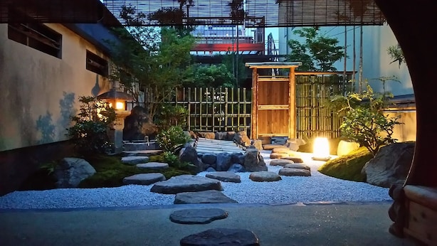 Un jardín japonés tradicional con un sendero de piedra que lleva a una puerta de madera, rodeado de rocas y follaje.