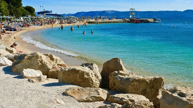 Una playa con aguas turquesas cristalinas, costa rocosa y un muelle con barcos.