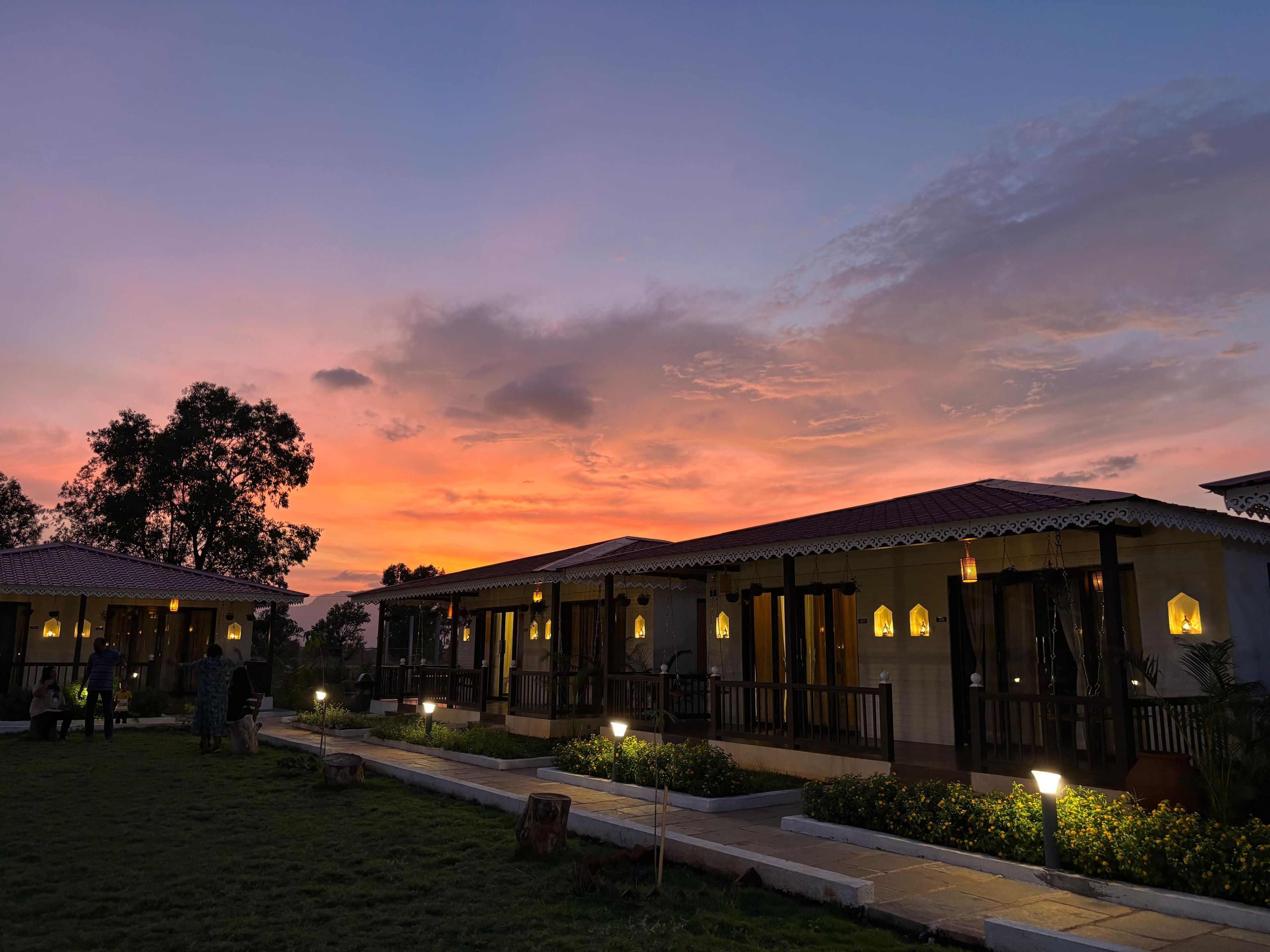 A row of traditional-style buildings with outdoor seating areas, connected by a pathway, and a sunset sky in the background.