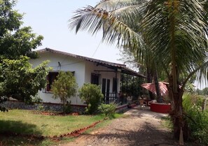 A house with a covered porch, a garden with various plants, and a dirt path leading to the entrance. - Mango Tree Farm (Karjat)