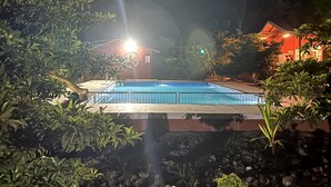 A nighttime view of a swimming pool surrounded by lush greenery and a building with a red roof. - Mango Tree Farm (Karjat)