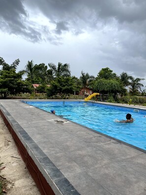 A swimming pool with a tiled edge, surrounded by greenery and a cloudy sky. - Mango Tree Farm (Karjat)