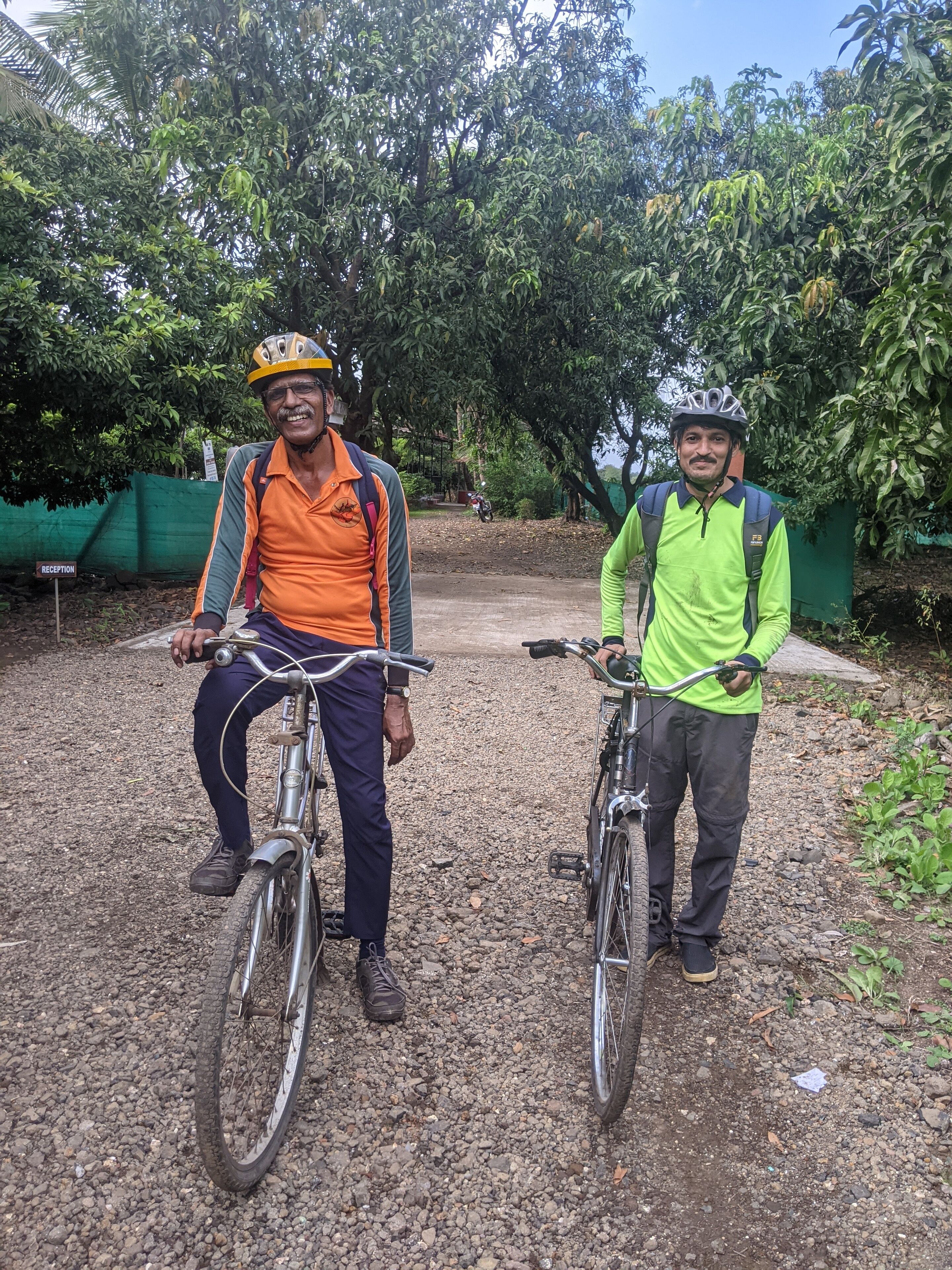 Dos personas en bicicleta, una vestida con una camiseta y casco naranja, y la otra con una camiseta y casco verdes, de pie sobre un sendero de grava con árboles al fondo.