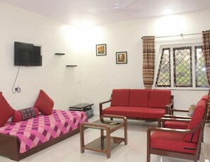 A living room with a sofa, armchairs, a coffee table, a TV, and a window with curtains. - Mango Tree Farm (Karjat)