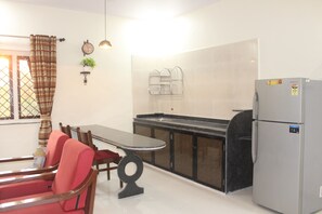 A compact kitchen with a dining area, a refrigerator, and a window with curtains. - Mango Tree Farm (Karjat)