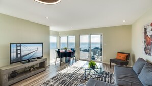A modern living room with a large TV, a sofa, and a dining area with a view of the ocean. - Otter Grotto (Victoria)