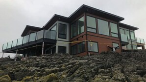 A modern two-story house with large windows and a balcony, built on a rocky hillside. - Otter Grotto (Victoria)