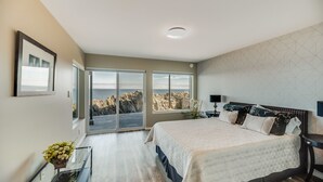 A bedroom with a large bed, a glass door leading to a balcony with a view of rocky terrain and the sea. - Otter Grotto (Victoria)