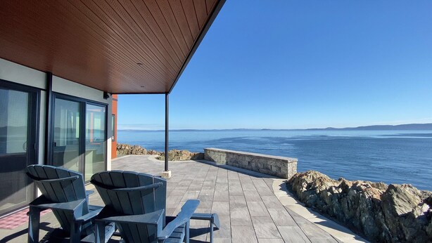 A covered outdoor seating area with chairs, a stone wall, and a view of the sea. - Otter Grotto (Victoria)