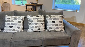 A grey sofa with patterned cushions, a magazine on a black coffee table, and a dining area with a table and chairs in the background. - Escape to Birchwood Suite (Fairbanks)