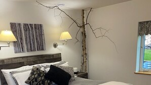 A bedroom with a bed, two bedside lamps, a nightstand, a window with a view, and a tree branch decoration on the wall. - Escape to Birchwood Suite (Fairbanks)