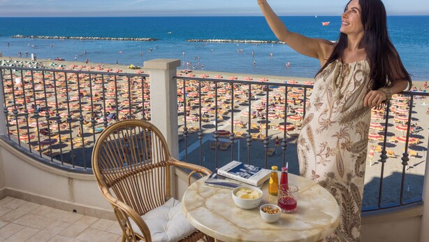 Un balcón con vista a una playa, una silla de mimbre, una mesa redonda con un libro y bebidas, y una barandilla.