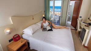 Habitación de hotel con una cama, una mesita de noche con un bolso rojo, una lámpara de lectura y vista a la playa a través de la puerta.