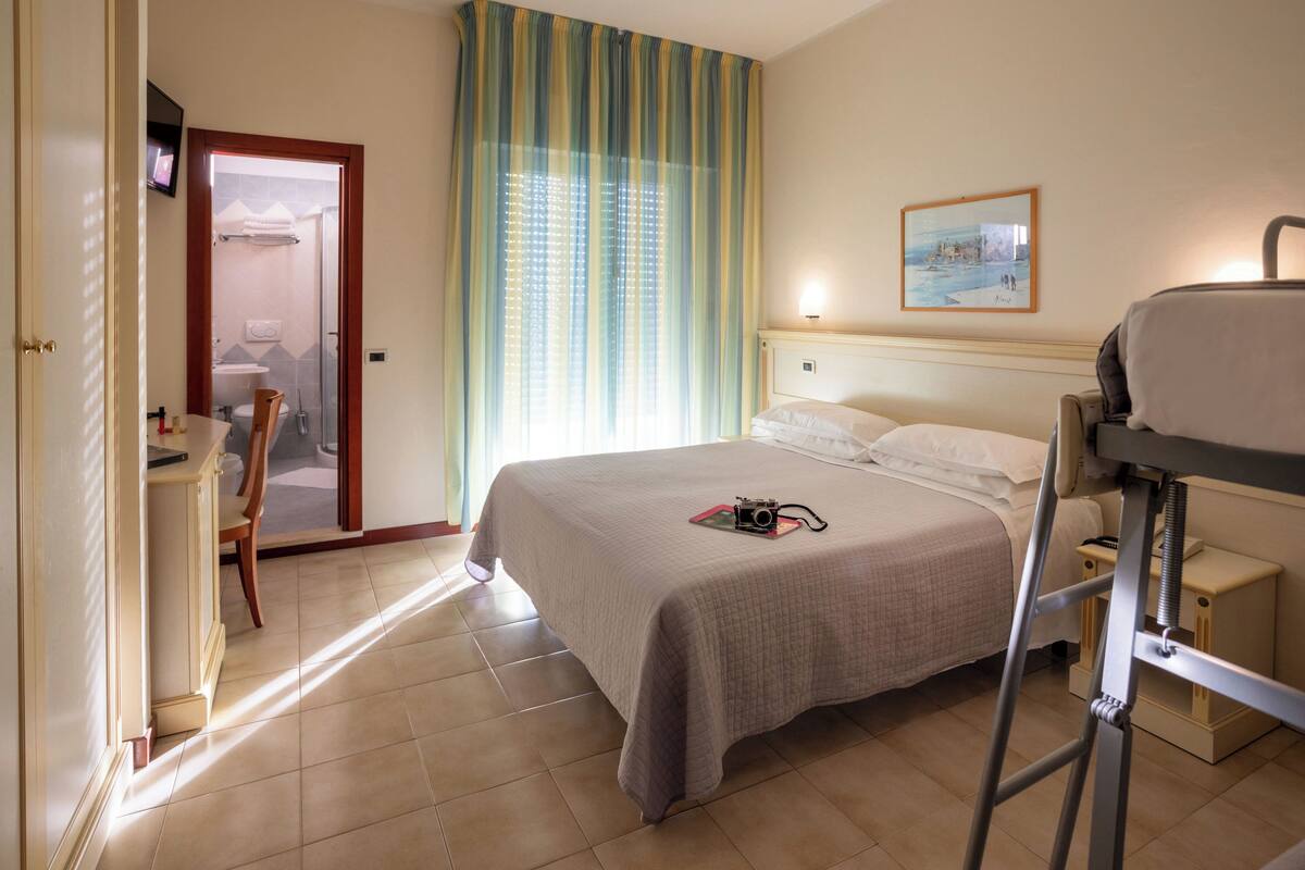 Habitación de hotel con cama, mesita de noche, silla y un baño visible a través de una puerta abierta.