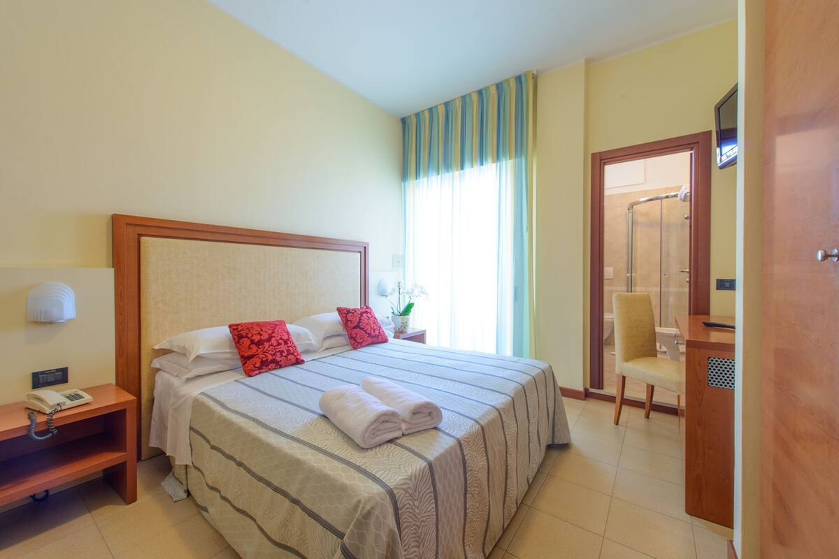 Habitación de hotel con una cama grande, una mesita de noche con teléfono, una silla y un escritorio con computadora.