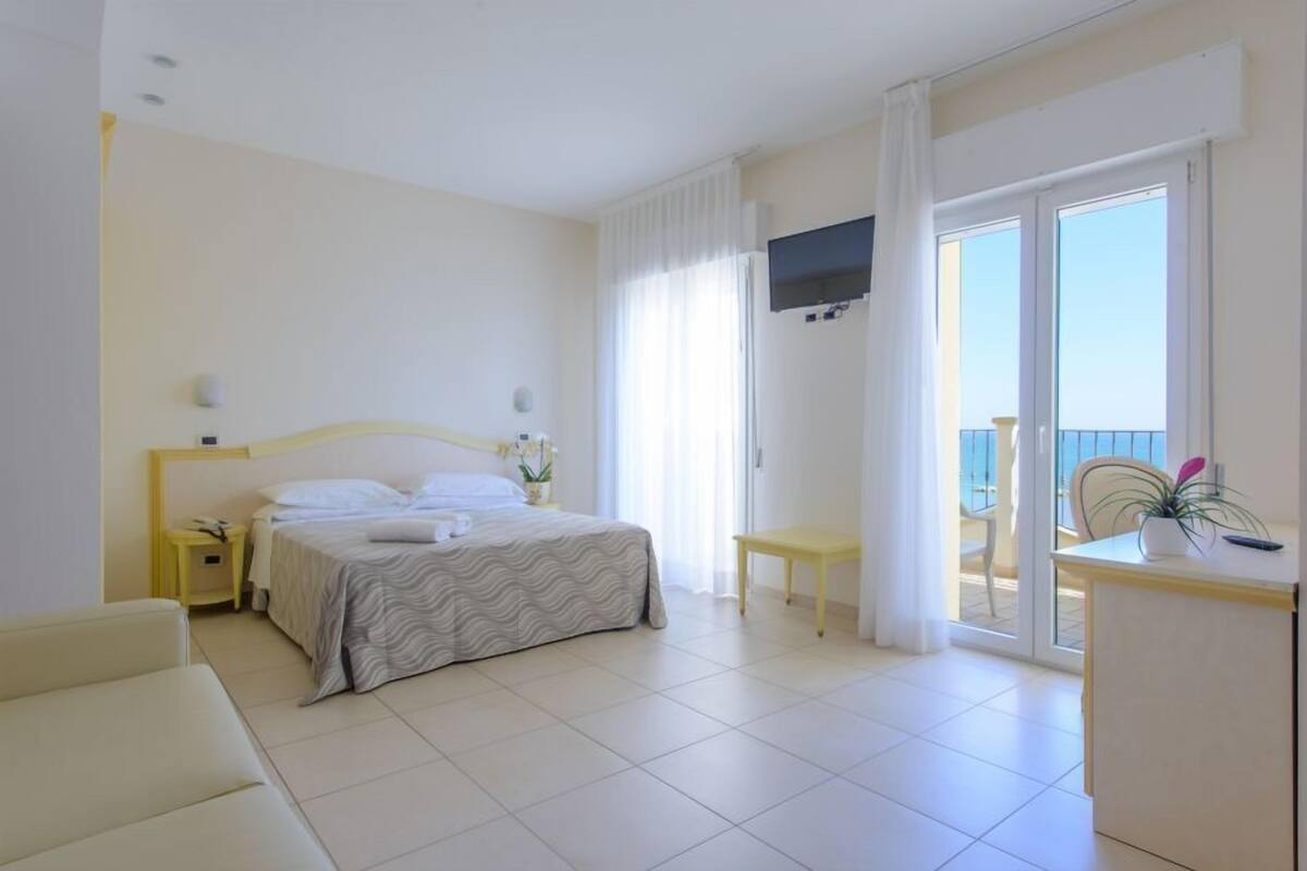 Habitación de hotel con cama, escritorio, silla, televisión y balcón con vistas.