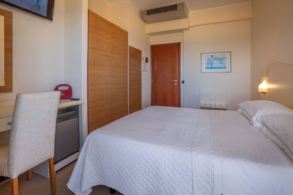 Una habitación de hotel con una cama, un escritorio con silla, un armario, una puerta, un aire acondicionado de pared y un cuadro enmarcado.