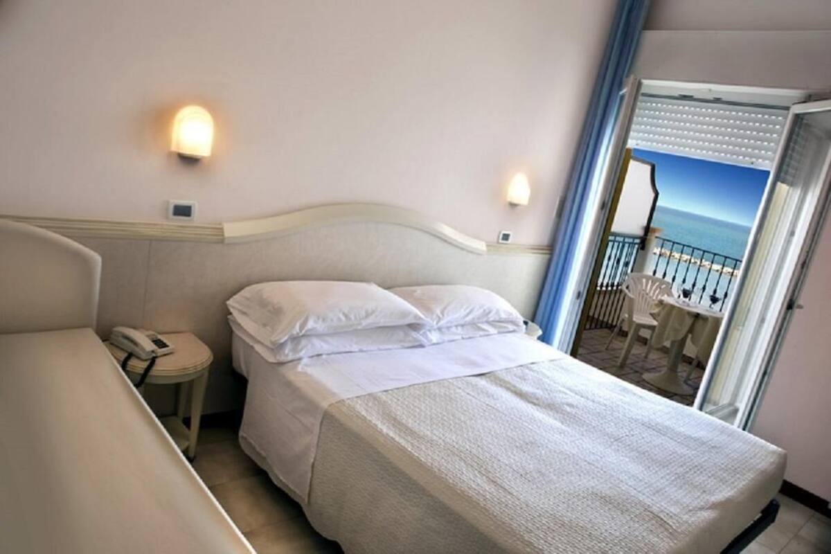 Habitación de hotel con cama, mesita de noche con teléfono y vista al mar a través de una puerta abierta.