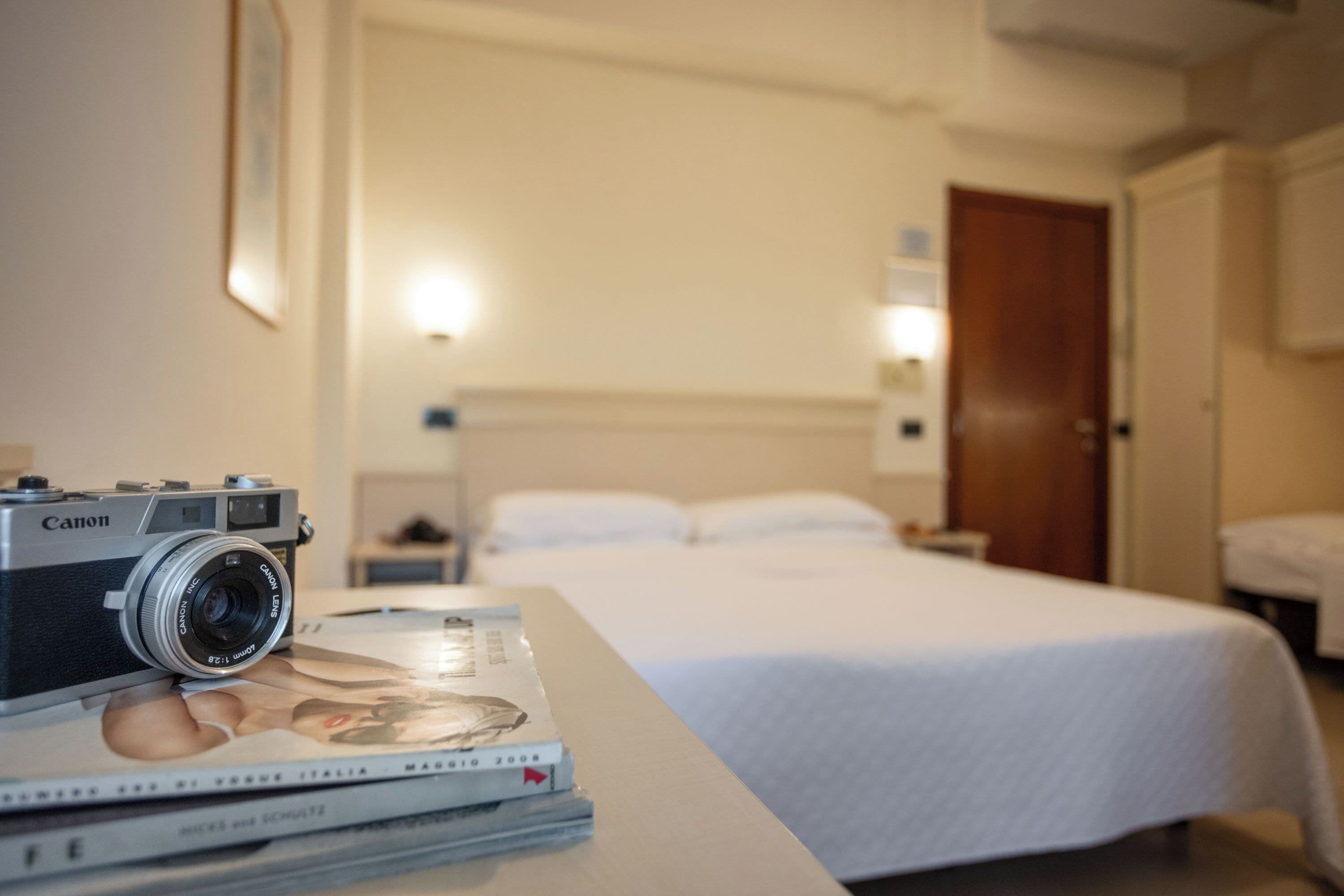 Une chambre d’hôtel avec un lit, une table de chevet sur laquelle se trouvent un appareil photo et des magazines, et une petite table de nuit.