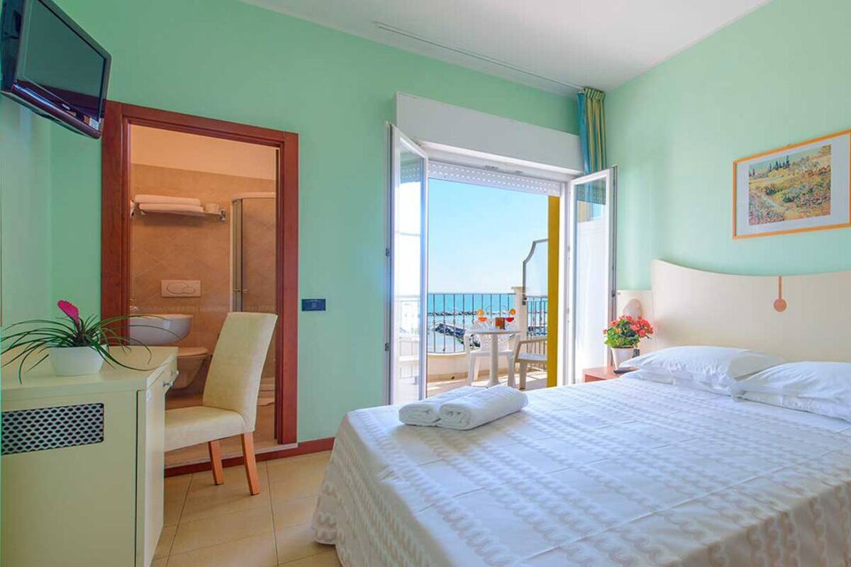 Habitación de hotel con cama, escritorio, silla, una mesita con planta y vista al balcón.