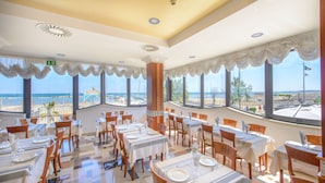 Un comedor con mesas dispuestas para el servicio, ventanas amplias con vistas a la playa y un techo con detalles decorativos.