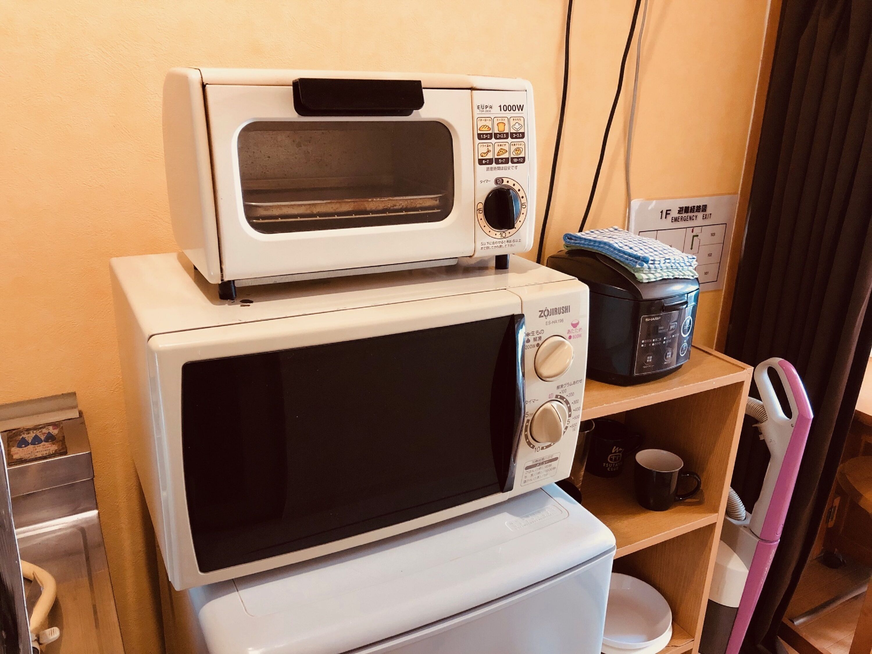 A kitchen with a white microwave oven stacked on top of a white toaster oven, a black rice cooker, and a small shelf with a blue cloth and a black mug.