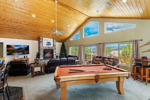A spacious living room with a pool table, leather sofas, a fireplace, and large windows offering views of the outdoors.