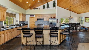 A spacious kitchen with wooden cabinets, a central island with bar stools, a stove, and a refrigerator. - Sky View Lodge by Destination Big Bear : Incredible Sky Valley Estates Offering Panoramic Meadow & Mountain Views! Spa! (Big Bear City)
