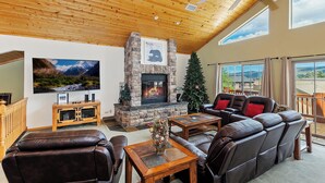 A spacious living room with a stone fireplace, leather sofas, a wooden coffee table, and a large TV on the wall. - Sky View Lodge by Destination Big Bear : Incredible Sky Valley Estates Offering Panoramic Meadow & Mountain Views! Spa! (Big Bear City)