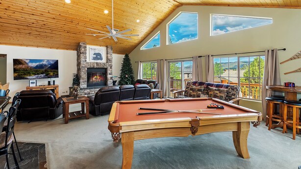 A spacious living room with a pool table, a fireplace, and large windows offering views of the outdoors. - Sky View Lodge by Destination Big Bear : Incredible Sky Valley Estates Offering Panoramic Meadow & Mountain Views! Spa! (Big Bear City)