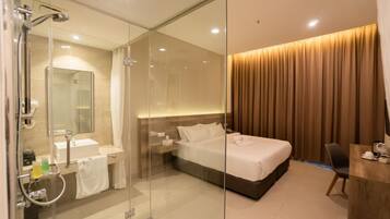 A modern hotel room with a glass-enclosed shower, a bed with a wooden headboard, a bedside table with a lamp, and a desk with a chair.