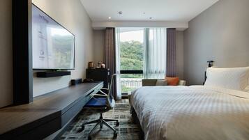 A modern hotel room with a large bed, a desk with a computer, a TV mounted on the wall, and a view of the outdoors.