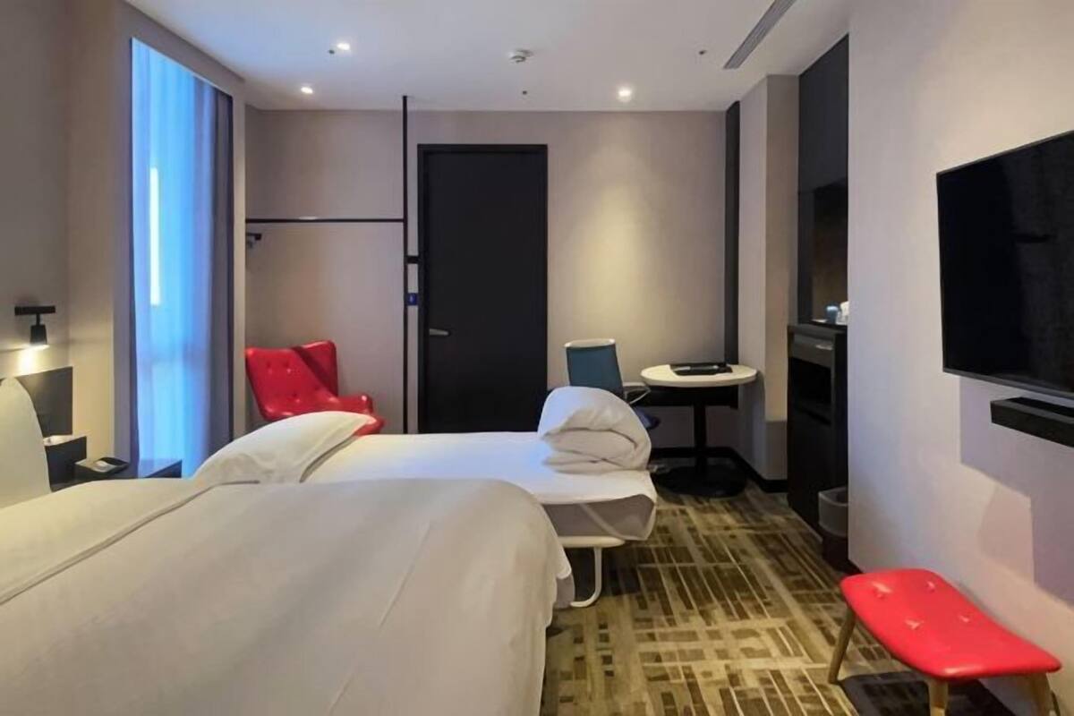 Ein Hotelzimmer mit zwei Betten, einem kleinen Tisch, einem Sessel, einem Fernseher und einem Fenster mit Vorhängen.