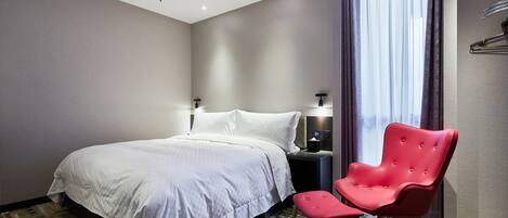 Quarto de hotel com cama, uma cadeira vermelha, mesa de cabeceira e janela com cortinas.