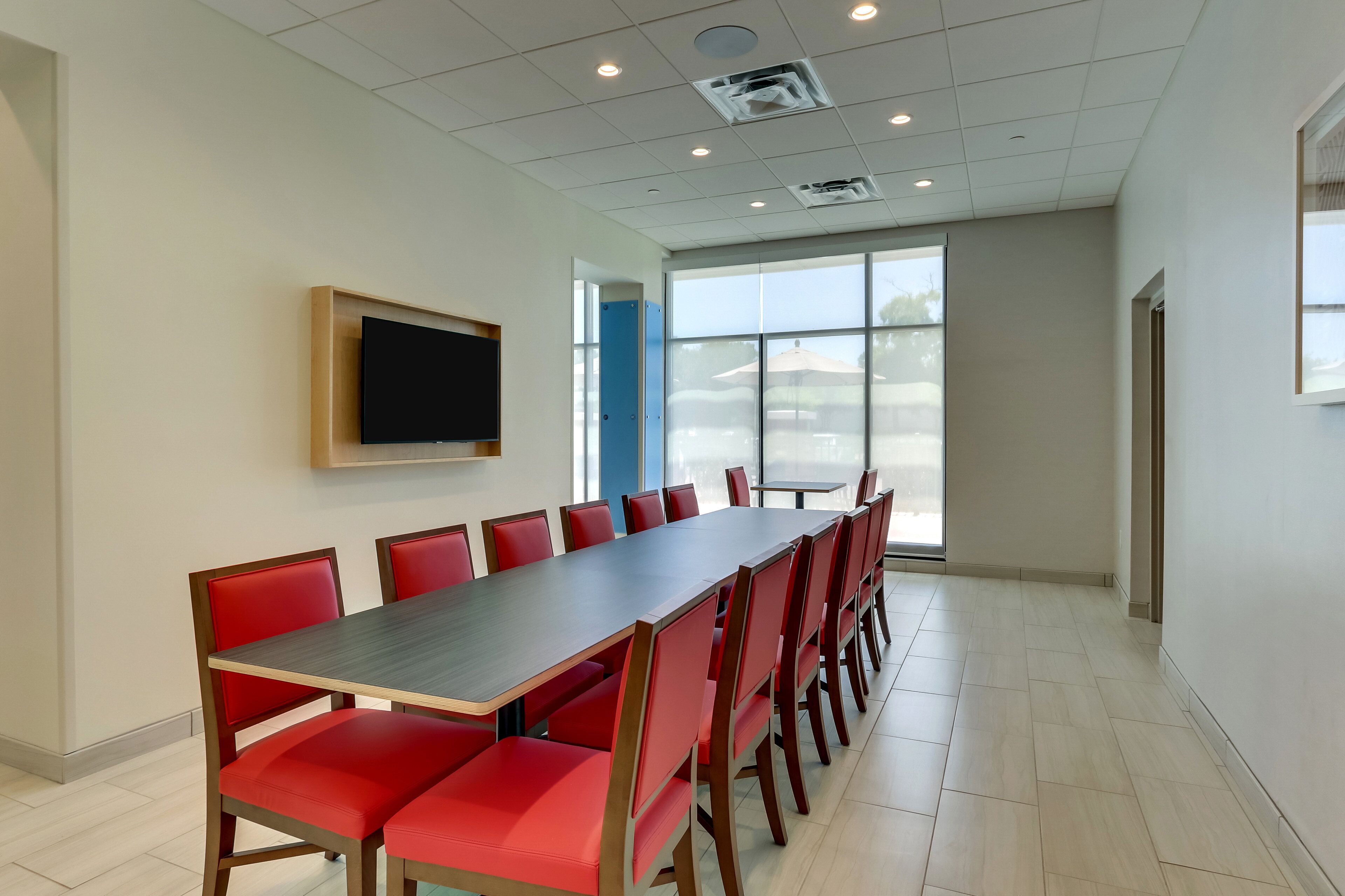 Un salón de conferencias con una larga mesa, sillas rojas, un televisor de pantalla plana y amplias ventanas.