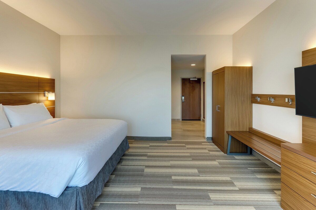 Una habitación de hotel moderna con una cama grande, cabecero de madera, un banco, un televisor y un armario.