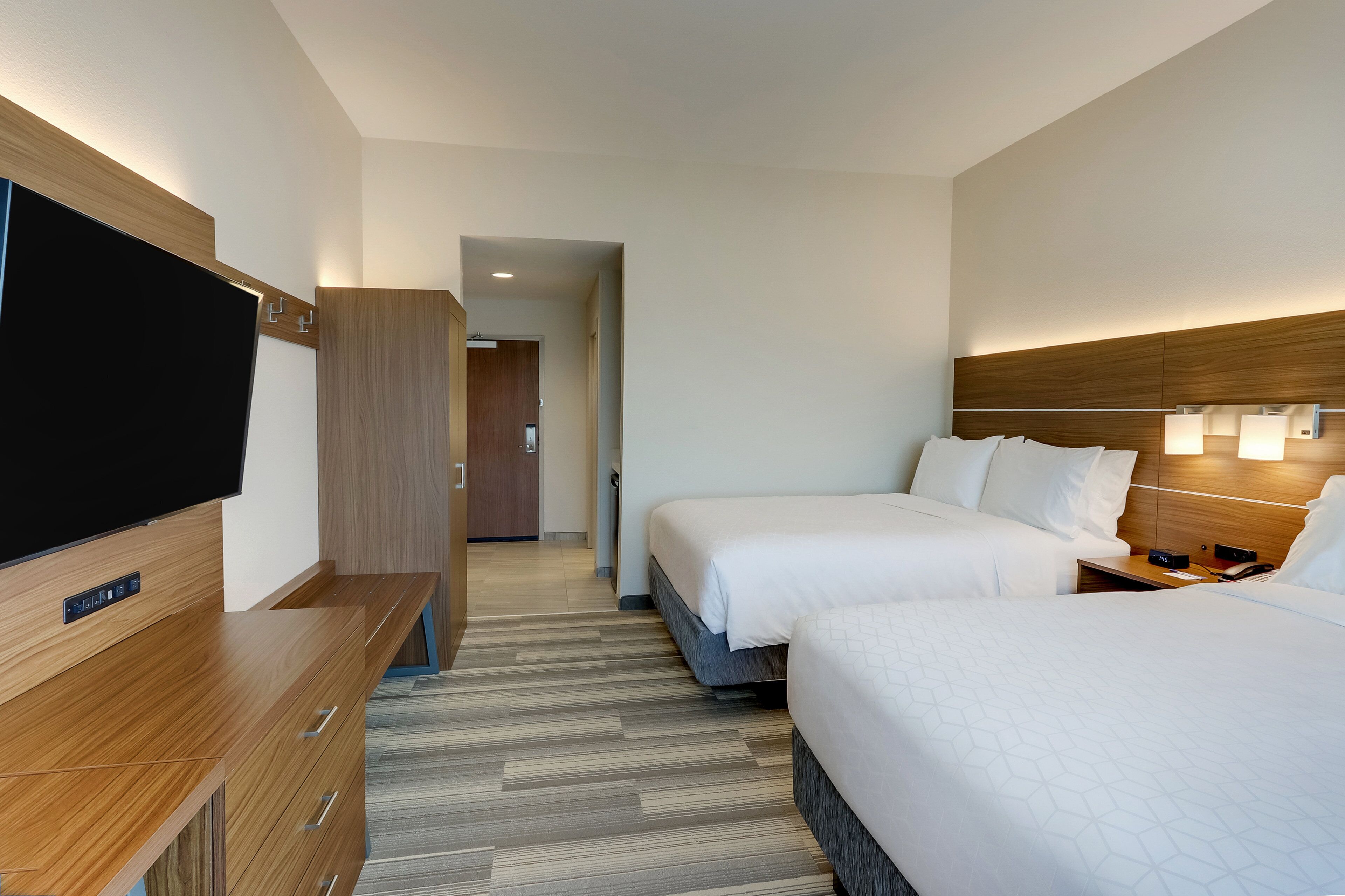A hotel room with a flat-screen TV, wooden furniture, two beds with white bedding, a wooden headboard, and a hallway leading to a bathroom.