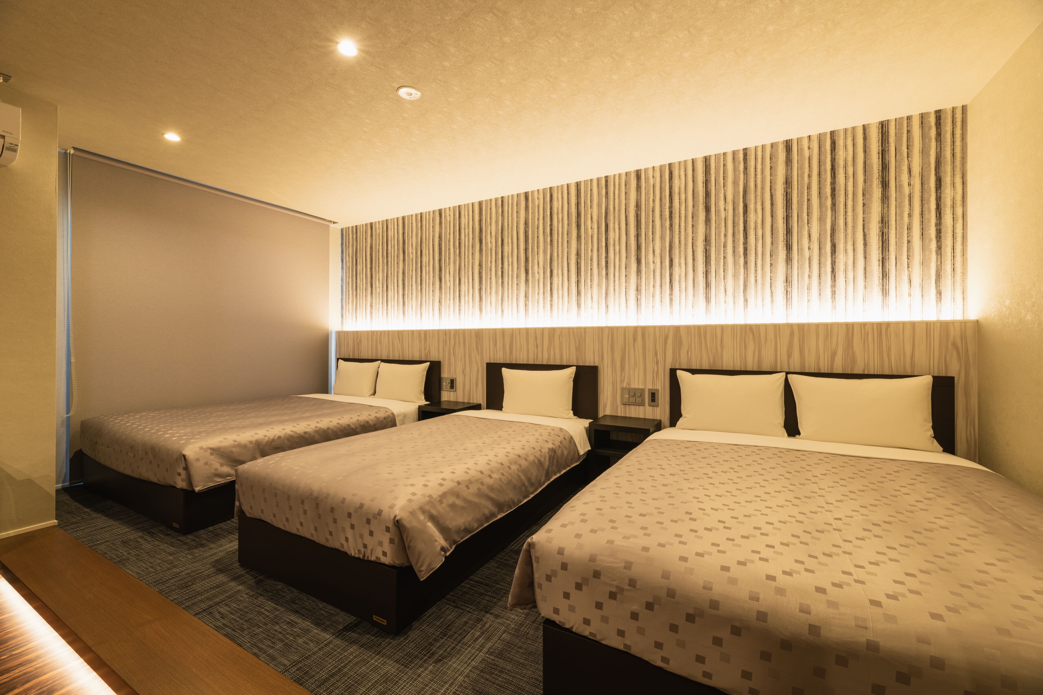 A hotel room with three single beds, a wooden bench, a large window, and a striped wall design.