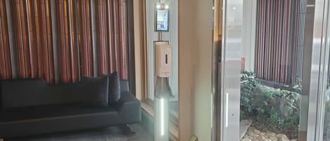 A reception area with a black sofa, a hand sanitizer dispenser, and a glass door leading outside.
