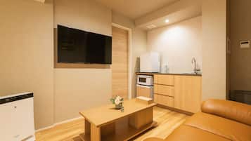 A compact living space with a sofa, a small wooden table, a flat-screen TV, a kitchenette with a microwave and refrigerator, and a compact washing machine.