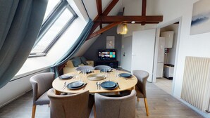 Ein Essbereich mit einem runden Tisch, gedeckt für sechs Personen, einem Fernseher im Hintergrund und Blick auf die Stadt durch ein großes Fenster.