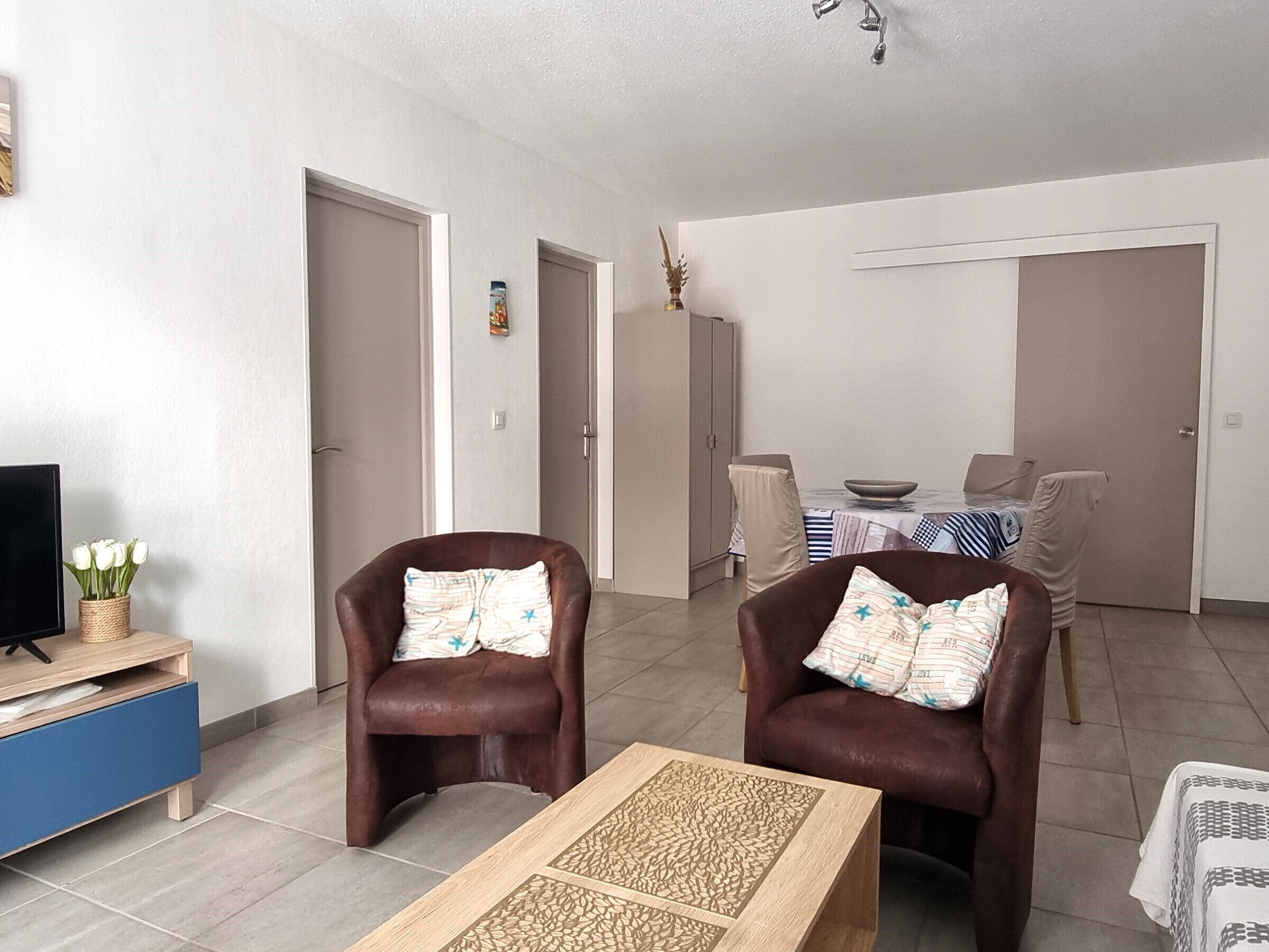 A living room with two brown leather chairs, a coffee table, a TV on a blue cabinet, and a dining area with a table and chairs.